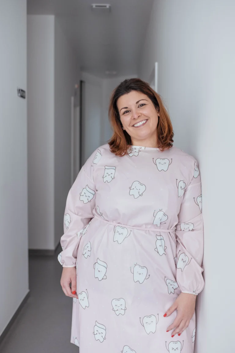 Mulher sorrindo em clínica odontológica usando avental com desenho de dentes. Ambiente moderno e limpo, transmitindo confiança e bem-estar na saúde oral.