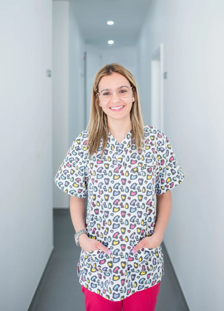 Mulher sorridente a usar uniforme, com jaleca colorida e calça rosa, no corredor da clínica, moderna com paredes brancas.