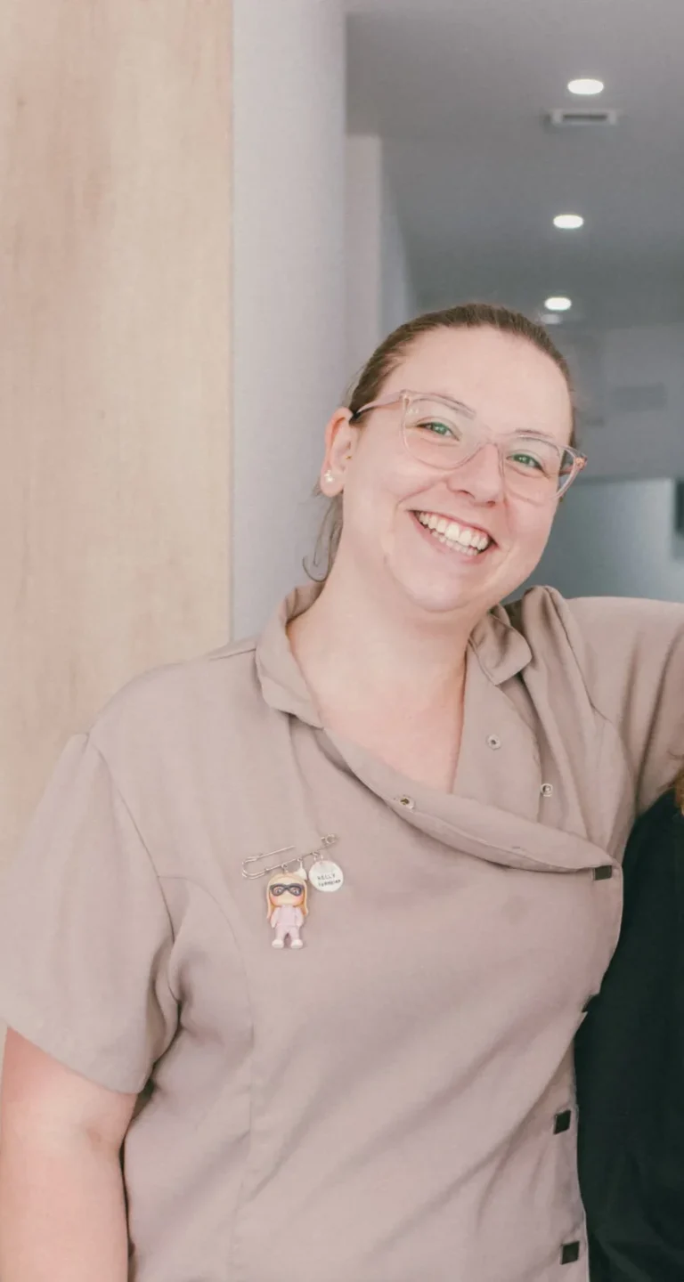 Mulher sorridente a usar uniforme casual em ambiente interno com parede branca ao fundo, transmitindo simpatia e profissionalismo, ideal para promover serviços de atendimento ao cliente ou equipe de trabalho.