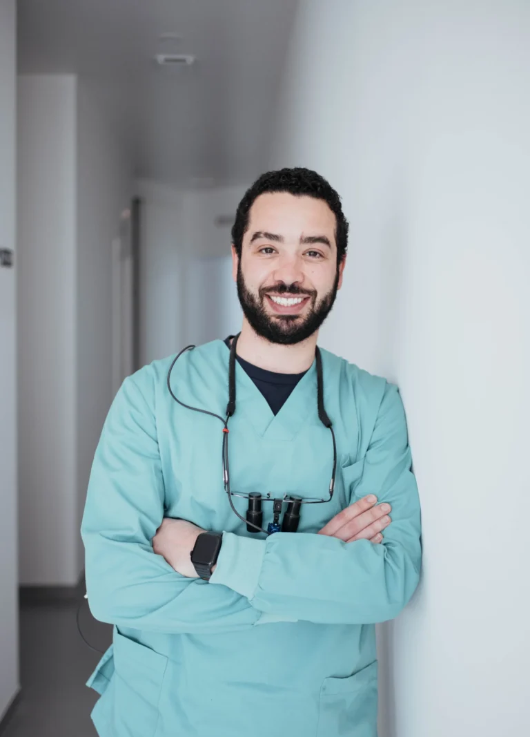 Médico sorridente, no corredor da clínica, a usar uniforme verde, com estetoscópio ao redor do pescoço, símbolo de cuidado e profissionalismo na área da saúde.