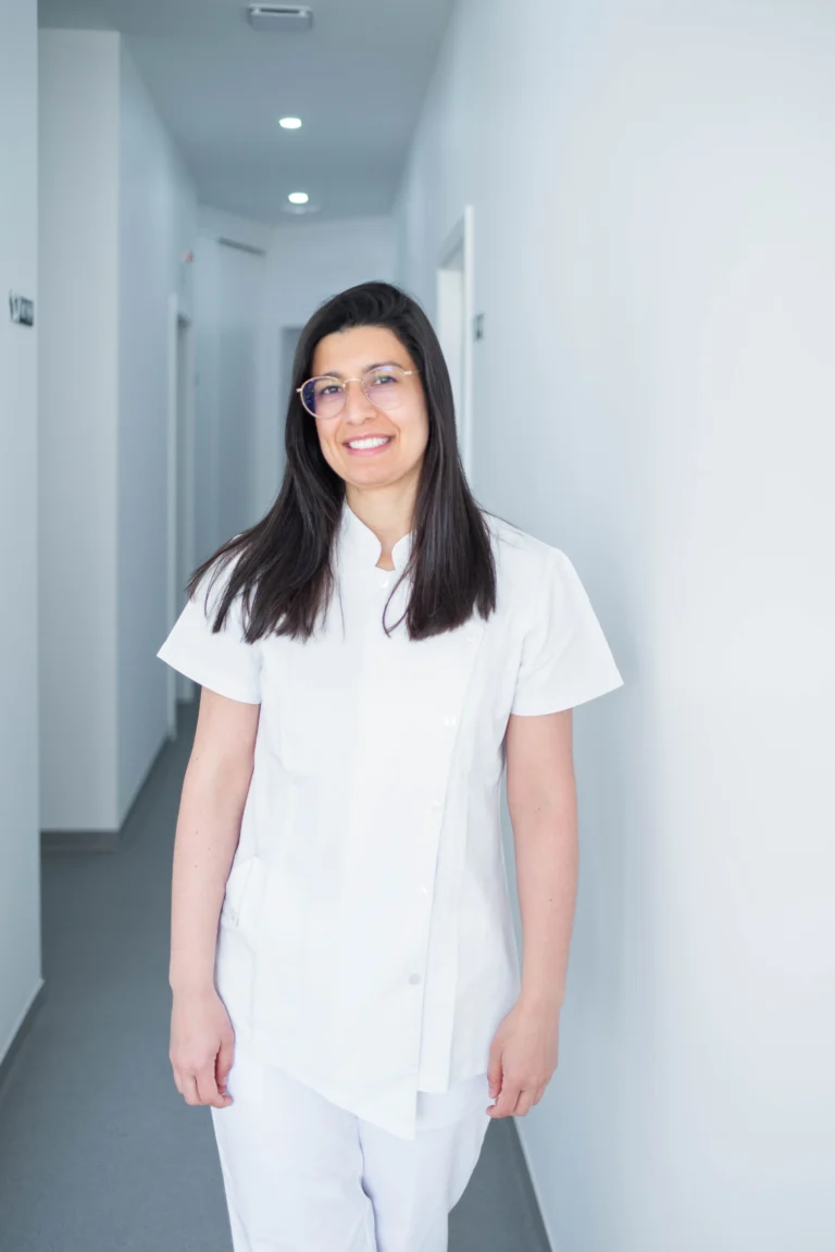 Profissional de saúde sorridente vestida com uniforme branco no corredor da clínica, representando cuidados médicos e bem-estar.