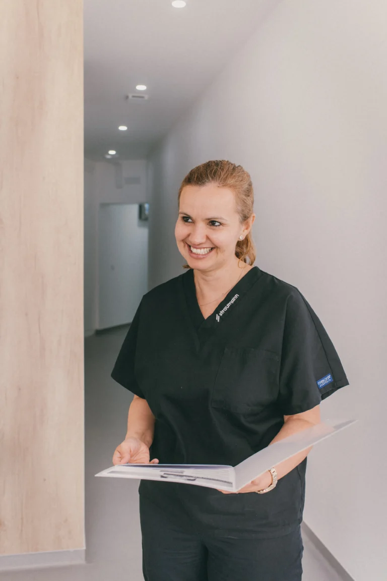 Profissional da área de saúde sorridente com uniforme preto, a segurar um prontuário, em ambiente clínico bem iluminado, transmitindo cuidado e atenção ao paciente.